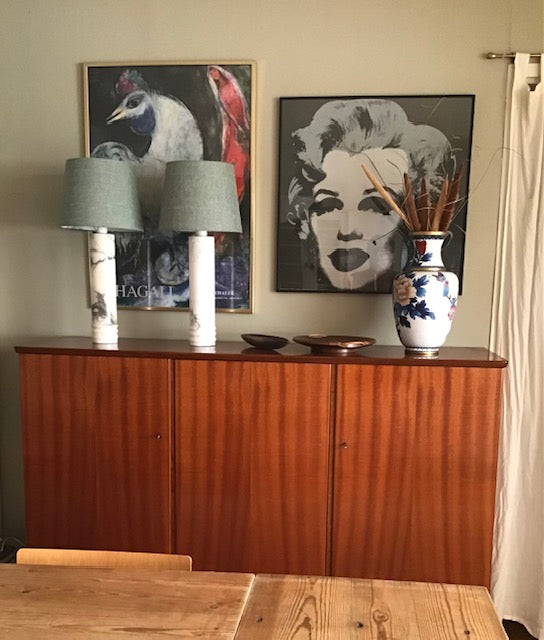 A mid-century modern wooden sideboard cabinet in a warm reddish-brown finish, styled with two gray lampshades on white marble bases, a blue and white floral vase, and two framed art prints on the wall behind it.