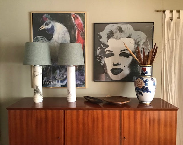 A styled interior scene showing a wooden sideboard with two table lamps, framed wall art including a Marilyn Monroe print, and a decorative ceramic vase with dried stems.
