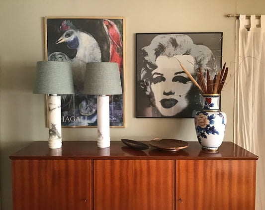 A styled interior scene showing a wooden sideboard with two table lamps, framed wall art including a Marilyn Monroe print, and a decorative ceramic vase with dried stems.