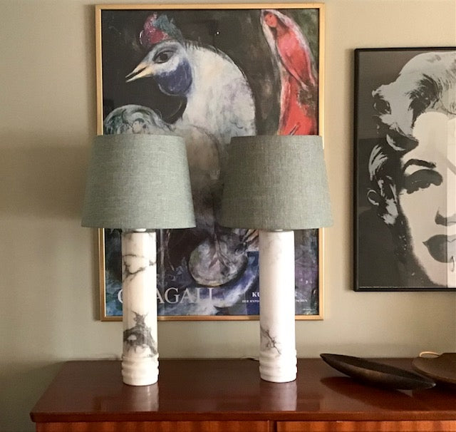 Two matching table lamps with gray fabric drum shades and white marble-patterned cylindrical bases, displayed on a wooden console table in front of framed wall art.