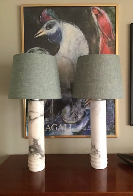 Two matching table lamps with gray fabric drum shades and white marble-look cylindrical bases featuring black veining, displayed on a wooden surface in front of a framed art print.