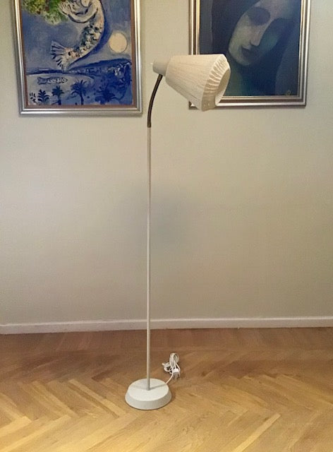 White floor lamp on a wooden floor with framed artwork on the wall.