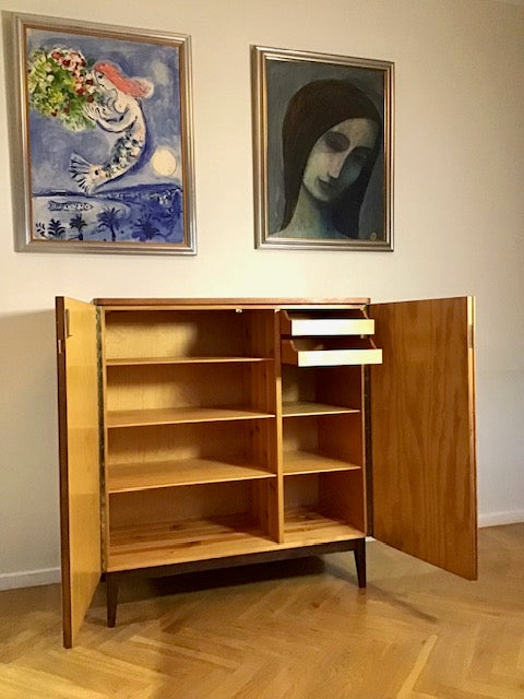 Wooden cabinet with open doors in a room with two framed artworks on the wall.